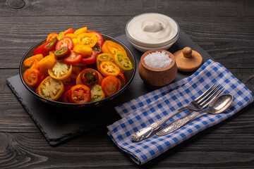 multicolored tomatoes cut into circles on a plate on a dark background with salt, sour cream and vegetable oil