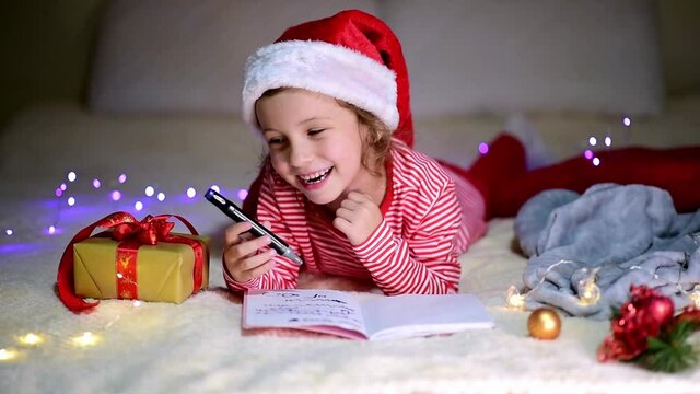 A little smiling girl in Santa's red hat and red christmas clothing lies on the bed and writes a letter to Santa Claus. New Year. hildren's Christmas. Atmosphere. Home.