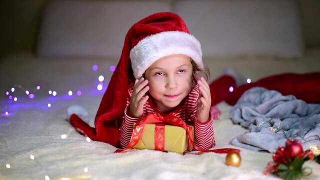 Children's Christmas. A little smiling girl in Santa's red hat and red christmas clothing lies on the bed with a gift. Preparation for the celebration. New Year. Atmosphere. Home.