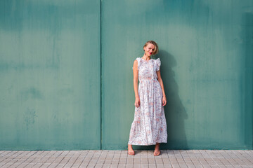 horizontal portrait of a blonde thin woman in floral summer dress. She has is leaning on a green wall.