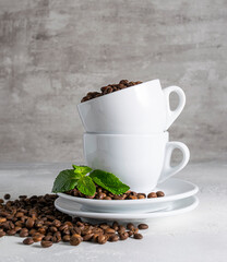 Fragrant coffee beans in a white cup. Coffee beans and mint leaves are on the saucer. Coffee beans are scattered on a light background