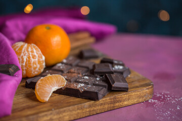 Close up of vegan dark chocolate bar and mandarin fruits on a wooden cutting board on pink table with dark background and holiday decorations