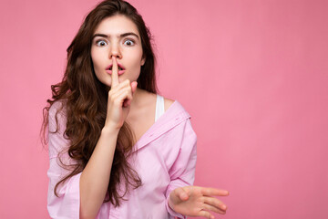 Photo of young shocked beautiful wavy-haired brunette woman with sincere emotions wearing casual pink shirt isolated over pink background with copy space and showing silence gesture with finger near