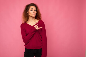 Young beautiful brunette wavy woman with sincere emotions wearing casual pink jersey isolated over pink background with copy space and pointing at free space for mockup