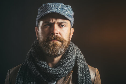 Portrait stylish brutal bearded man wears hat, scarf&leather jacket. Bearded hipster man dressed in jacket and wool cap. Guy with beard and mustache in tweed cap&leather coat. Macho in stylish clothes