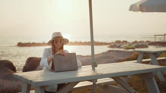 Pretty caucasian woman in sunglasses and hat sitting at wooden table and working on laptop. Sunshine over sea on background. Freelance and travelling concept.
