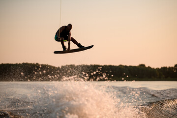 muscular man jumping high with wakeboard over splashing water against the backdrop of sky