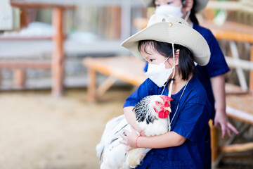 Side view of a cute girl wearing a white 4D shaped face mask. Little child wears a cream colored hat. Kid is holding or hugging the chicken to her body. Sweet smile baby.