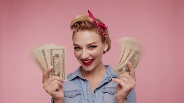 Attractive young retro female holding cash dollars in hands. Caucasian girl hold fan of cash money in dollars banknotes. Woman posing isolated on pink background studio with cash.