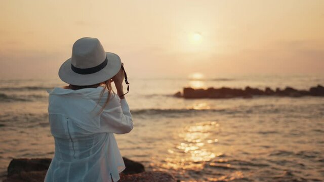 Happy female traveler in white shirt and hat standing on seaside and taking picture of beautiful summer sunset. Caucasian woman using digital camera for photographing.