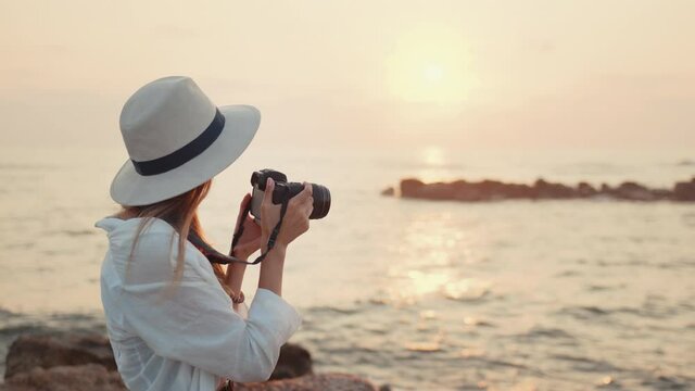 Back view of young woman in summer clothes and hat using modern camera for taking photos of amazing sunset over sea. Concept people, lifestyles and summertime.