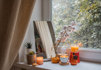 Autumn composition of candles, mirror, flowers and pumpkin figurines by the window in girl's room....
