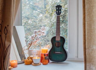 Autumn composition of candles, mirror, flowers, pumpkin figurines and ukulele on the windowsill in...