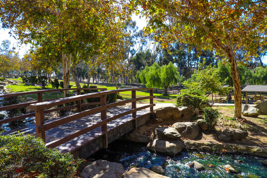 a gorgeous shot of an arched wooden bridge over a river with water rushing over rocks surrounded by lush green and autumn colored trees and blue sky at Kenneth Hahn Recreation Area in Los Angeles CA