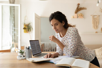 Focused adult female student doing homework, reading books at laptop, taking notes. New mother learning online from home, getting new occupation, preparing for college test. Education concept