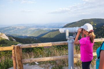 Fototapeta na wymiar Little girl hiker, observing nature landscape through stationary binocular telescope on mountain peak.