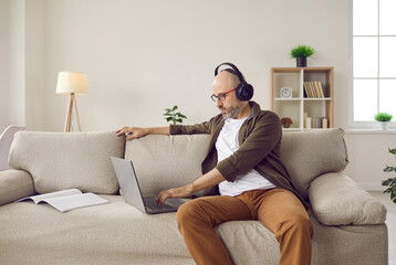 Adult relaxed man listening to music on headphones while working on laptop at home. Middle-aged Caucasian man sits next to laptop and notebook on comfortable sofa in cozy living room.