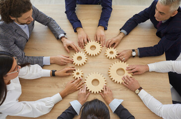 Team of businesspeople developing good business system. Group of senior and young business people sitting around table join gearwheels as metaphor for good effective teamwork. High angle, from above - obrazy, fototapety, plakaty