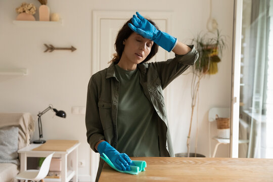 Tired Overheated Housewife Doing Domestic Work At Home, Cleaning Apartment. Exhausted Woman Wearing Protective Gloves, Dusting Furniture, Suffering From Heat, Hot Air. Household, Overwork Concept