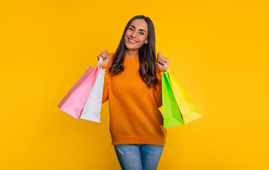 Fototapeta na wymiar Cute fashionable young smiling woman holds many colorful shopping paper bags in hand while she is isolated on yellow background