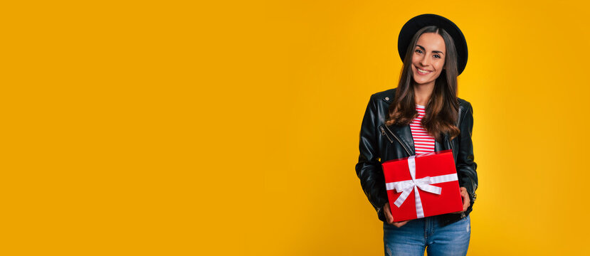 Wide-length banner photo of fashionable hipster smiling woman with big red present box in hands while she posing in studio with yellow wall