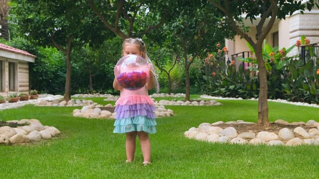 funny little girl, 3 years old, with two ponytails on her head, dressed in a delicate and multi-colored dress of pink blue color, plays with a bright transparent ball with multi-colored feathers