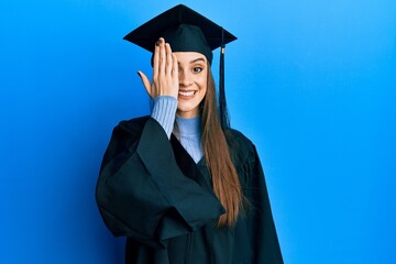 Beautiful brunette young woman wearing graduation cap and ceremony robe covering one eye with hand, confident smile on face and surprise emotion.