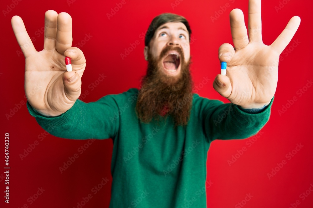 Poster redhead man with long beard holding two different pills angry and mad screaming frustrated and furio