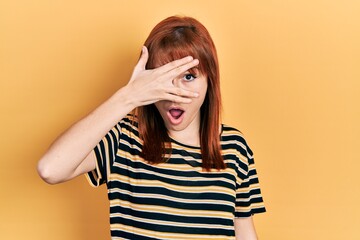 Redhead young woman wearing casual striped t shirt peeking in shock covering face and eyes with hand, looking through fingers afraid