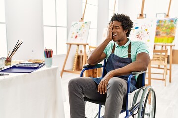 Young african american artist man sitting on wheelchair at art studio covering one eye with hand,...