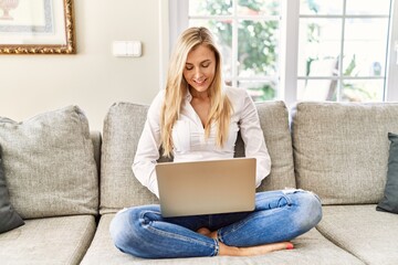 Young blonde woman using laptop sitting on the sofa at home.