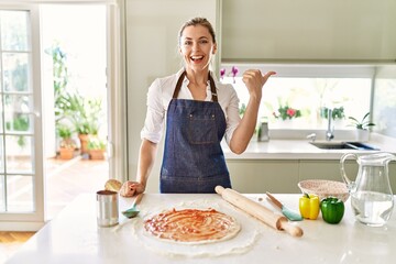 Beautiful blonde woman wearing apron cooking pizza smiling with happy face looking and pointing to the side with thumb up.