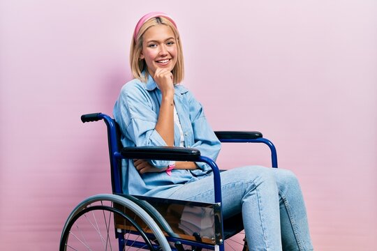 Beautiful blonde woman sitting on wheelchair looking confident at the camera smiling with crossed arms and hand raised on chin. thinking positive.
