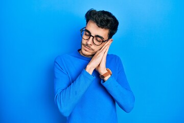 Young hispanic man wearing casual clothes and glasses sleeping tired dreaming and posing with hands together while smiling with closed eyes.