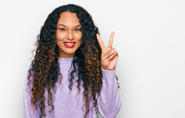 Young hispanic woman with curly hair wearing casual winter sweater smiling with happy face winking at the camera doing victory sign. number two.