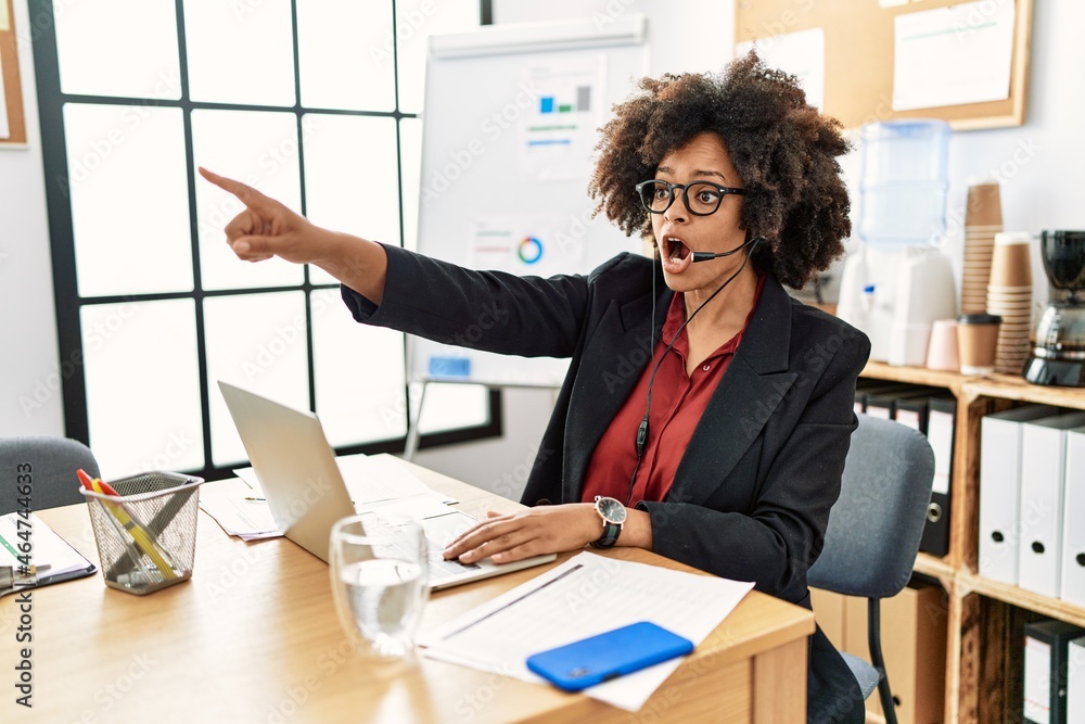 Wall mural African american woman with afro hair working at the office wearing operator headset pointing with finger surprised ahead, open mouth amazed expression, something on the front