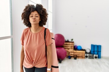 African american woman with afro hair holding yoga mat at pilates room looking at the camera blowing a kiss on air being lovely and sexy. love expression.