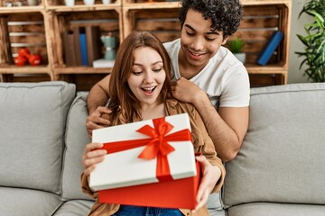 Young couple smiling happy sitting on the sofa surprise with anniversary gift at home.