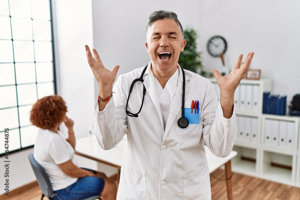 Sticker Middle age doctor man at the clinic with a patient celebrating mad and crazy for success with arms raised and closed eyes screaming excited. winner concept