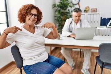 Senior woman sitting at doctor appointment looking confident with smile on face, pointing oneself with fingers proud and happy.