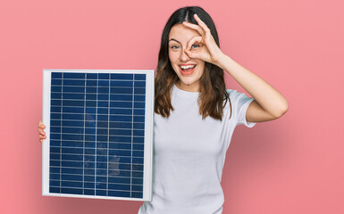 Young beautiful girl holding photovoltaic solar panel smiling happy doing ok sign with hand on eye...