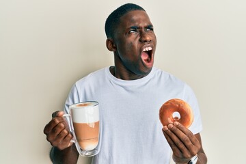 Young african american man eating doughnut and drinking coffee angry and mad screaming frustrated and furious, shouting with anger. rage and aggressive concept.