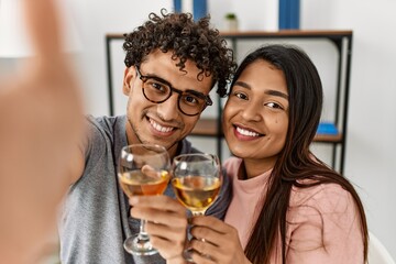 Young latin couple make selfie by the camera toasting with wine sitting on the table at home.