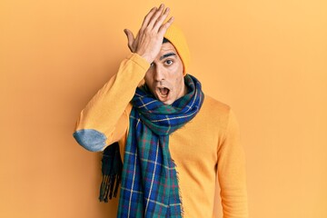 Handsome hispanic man wearing wool hat and winter scarf surprised with hand on head for mistake,...