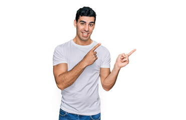 Handsome hispanic man wearing casual white t shirt smiling and looking at the camera pointing with two hands and fingers to the side.