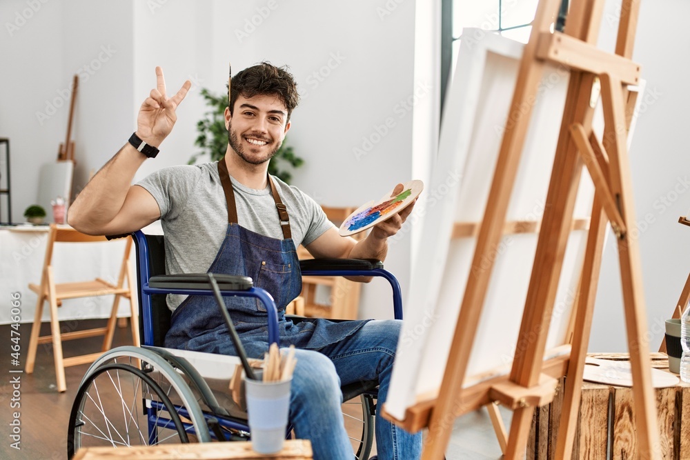 Poster young hispanic man sitting on wheelchair painting at art studio smiling with happy face winking at t