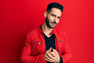 Young hispanic man wearing red leather jacket with hands together and crossed fingers smiling relaxed and cheerful. success and optimistic
