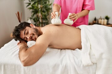 Obraz na płótnie Canvas Young hispanic man smiling happy reciving herbal pouches thai massage at beauty center.