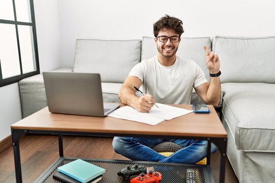 Hispanic man doing papers at home gesturing finger crossed smiling with hope and eyes closed. luck and superstitious concept.