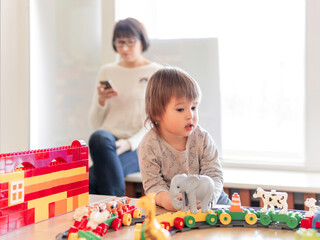 Toddler plays with colorful toy blocks while his mother or babysitter texting in smartphone. Little boy stares on toy constructor. Interior of kindergarten or nursery.
